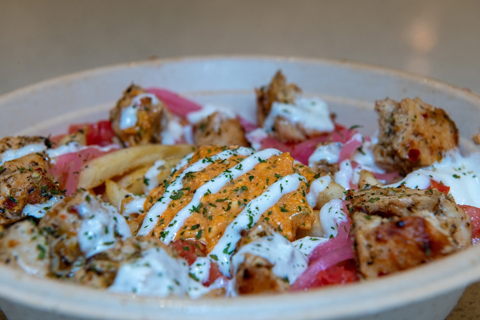 A close-up of a Mediterranean bowl from Luna Grill featuring grilled chicken, crispy fries, pickled onions, creamy tzatziki, and spicy feta sauce, topped with fresh herbs.