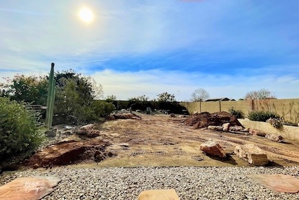 A sunny Arizona backyard undergoing a native landscaping transformation, with cleared soil, large desert rocks, surrounding shrubs, and a bright blue sky overhead.